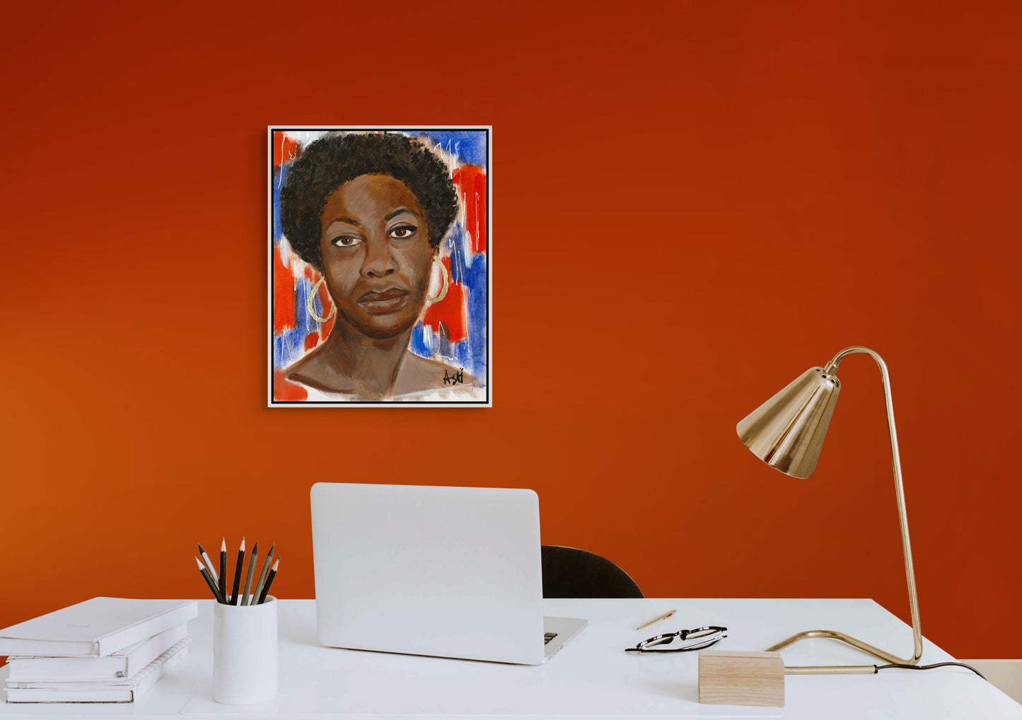 Framed stretched canvas print of 'Red, White & Blues - Nina Simone,' displayed in a white frame on a bold red wall above a desk. The artwork features Simone against a vibrant red, white, and blue background, celebrating her impact in music and civil rights, with the setting adding a striking contrast.