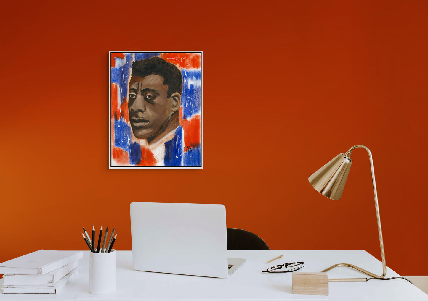 Framed stretched canvas print of 'Red, White & Blues - James Baldwin,' elegantly framed in white and hung on a striking red wall above a desk. The artwork features Baldwin against a dynamic red, white, and blue background, celebrating his legacy in literature and civil rights, with the setting providing a bold contrast.