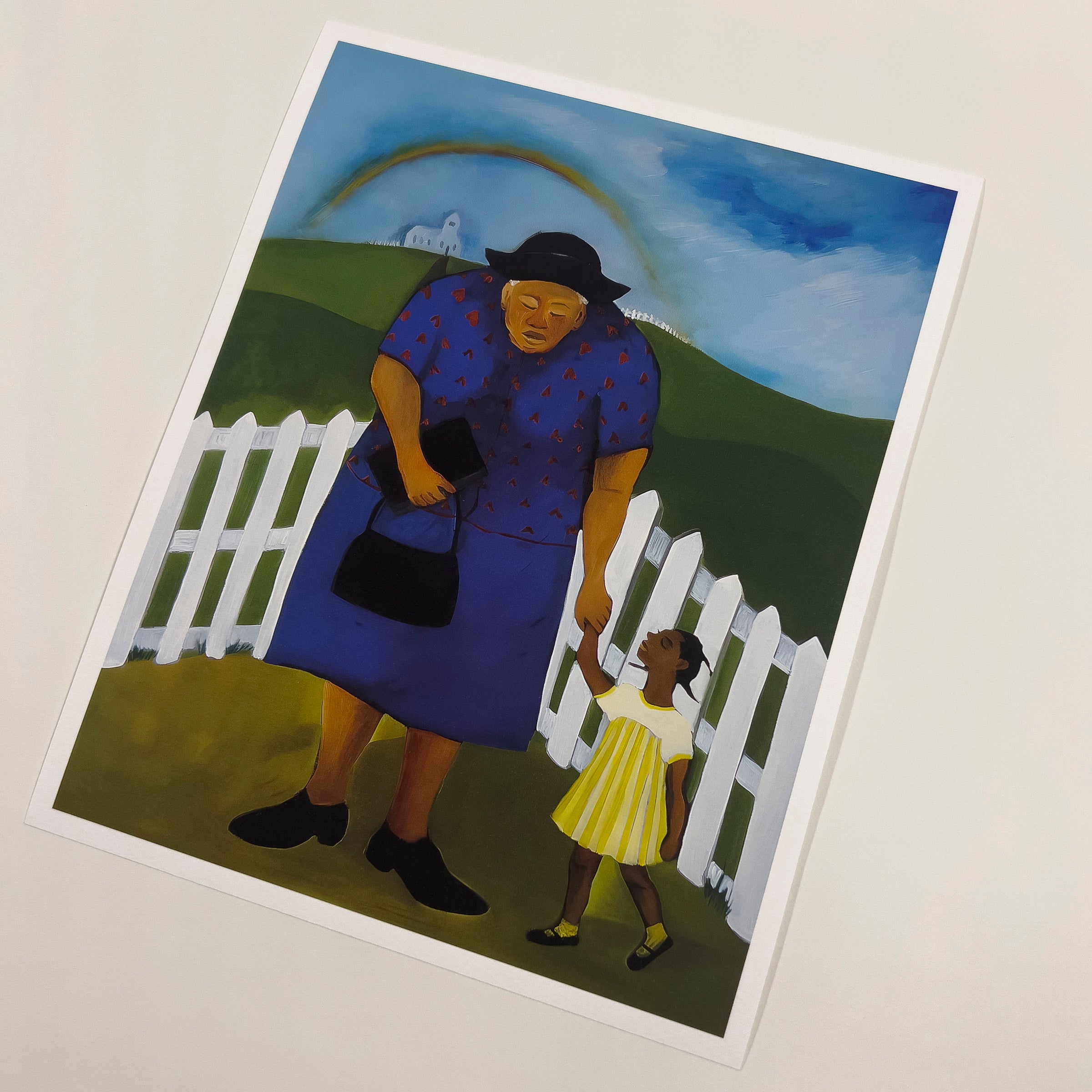 An art print showing a young Black girl in a yellow dress walking hand in hand with her elder toward a white church on a hill, beneath a bright rainbow and blue sky, surrounded by green fields and a white picket fence.